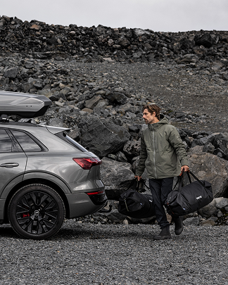 A man carrying two black Thule duffel bags approaches a car with a Thule roofbox mounted to the top against a rocky background.