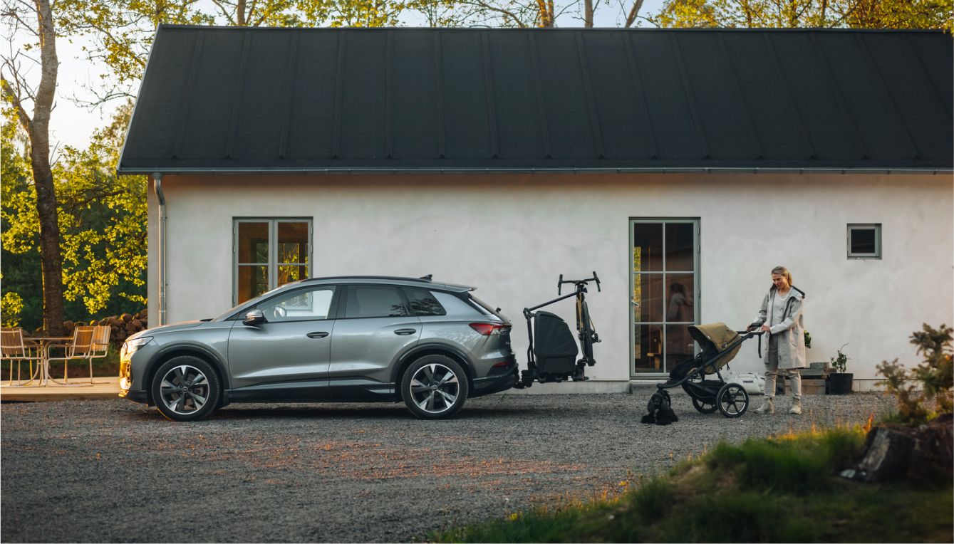 一名女子站在一栋现代住宅附近的婴儿车和小狗旁，旁边的碎石车道上停着一辆配备 Thule Onto 2 后置载物箱和自行车架的灰色汽车。