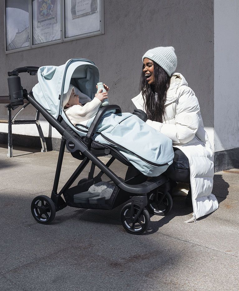 A woman kneels down to her child in a stroller with a footmuff.