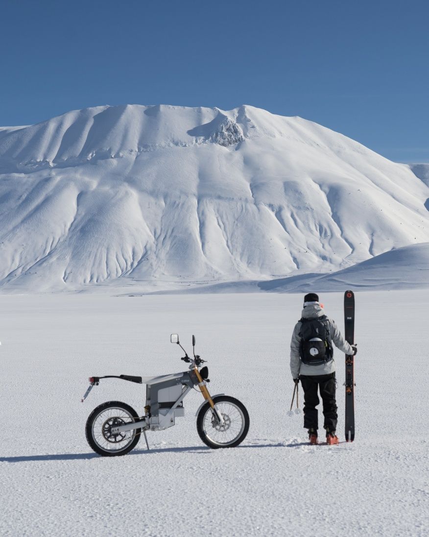 A man stands next to a motorbike while holding a pair of skis on the snow.