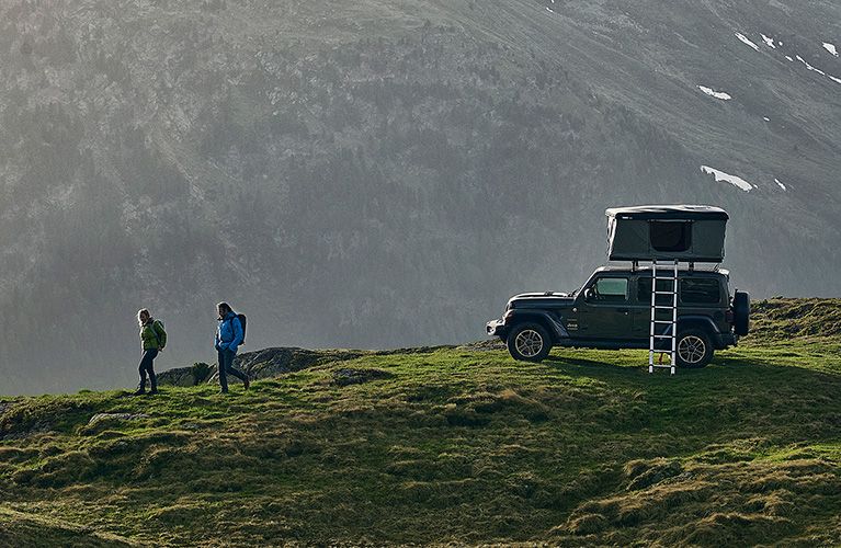 Two hikers walk by a mountain where a jeep is parked with a hard-shell rooftop tent.