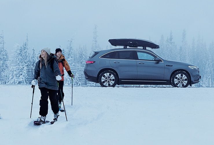 Två skidåkare ger sig ut i spåret och deras elbil med skidbox på taket står i bakgrunden.