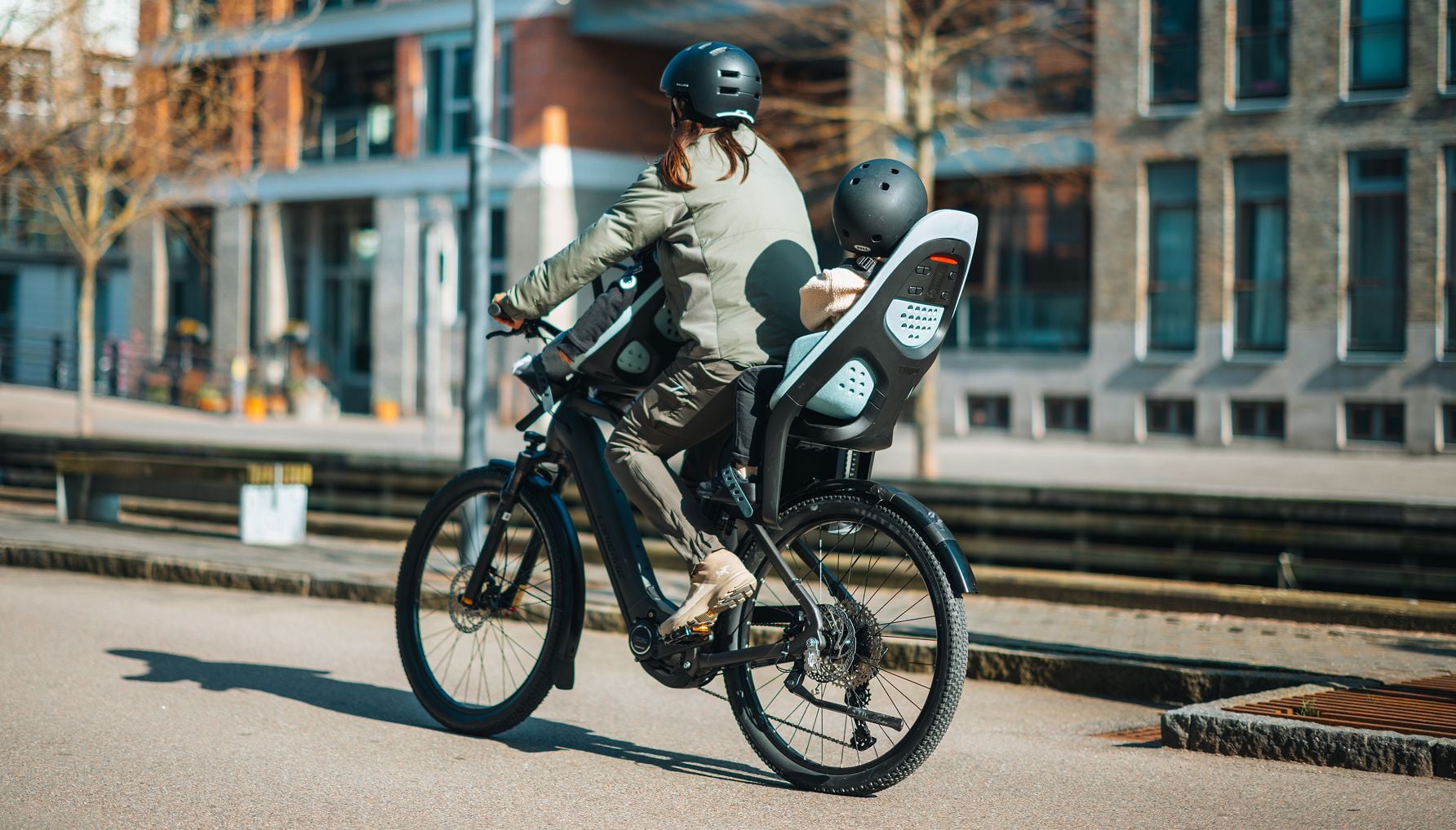 Adult fastening a child’s harness straps while the child sits in a rear bike seat, both wearing helmets.