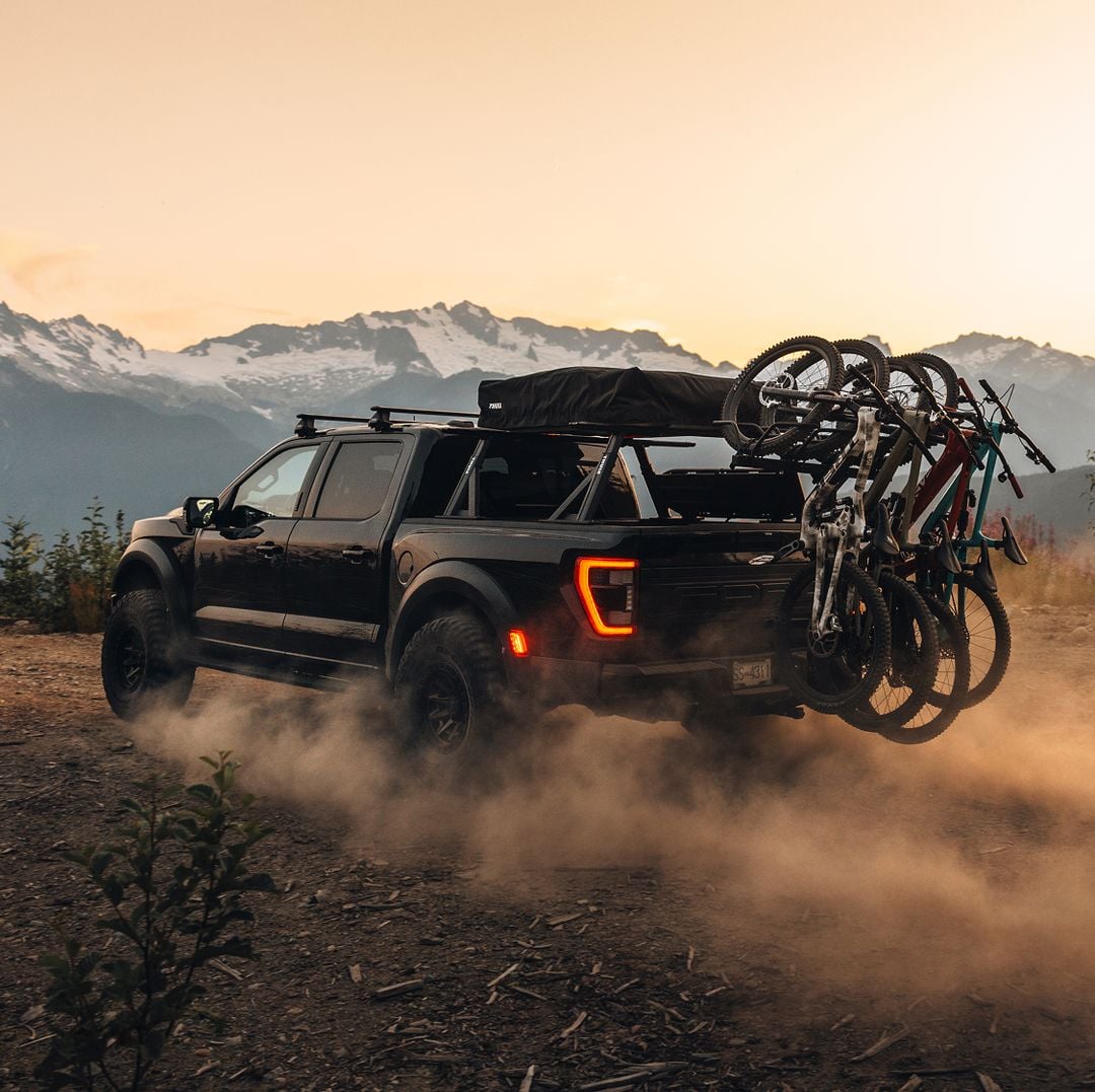 Pickup truck driving on a dusty mountain road at sunset with multiple bikes mounted on a Thule Revert bike rack, promoting Bring Your Bike.