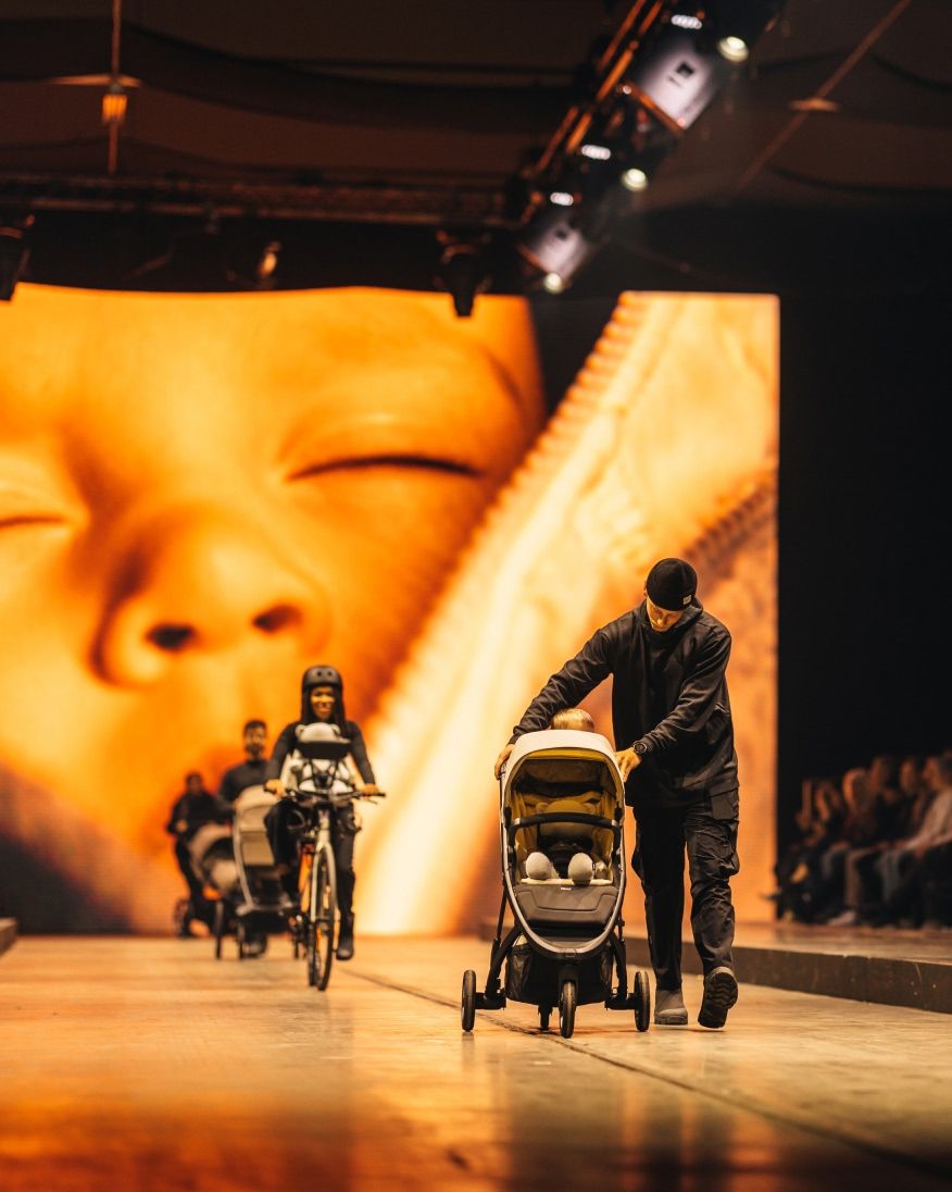 A person pushing a stroller on a runway during a show, with a large close-up image of a baby displayed on the screen behind them.