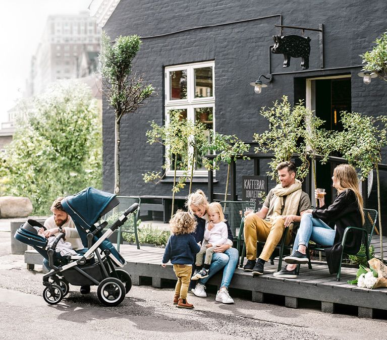 Parents and kids outside a cafe playing and drinking juice, a father tends to a baby in a twin stroller.