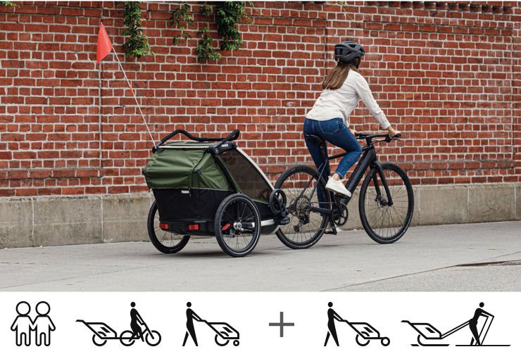 A woman bikes past a brick wall with her green Thule Chariot Cab baby bike trailer.