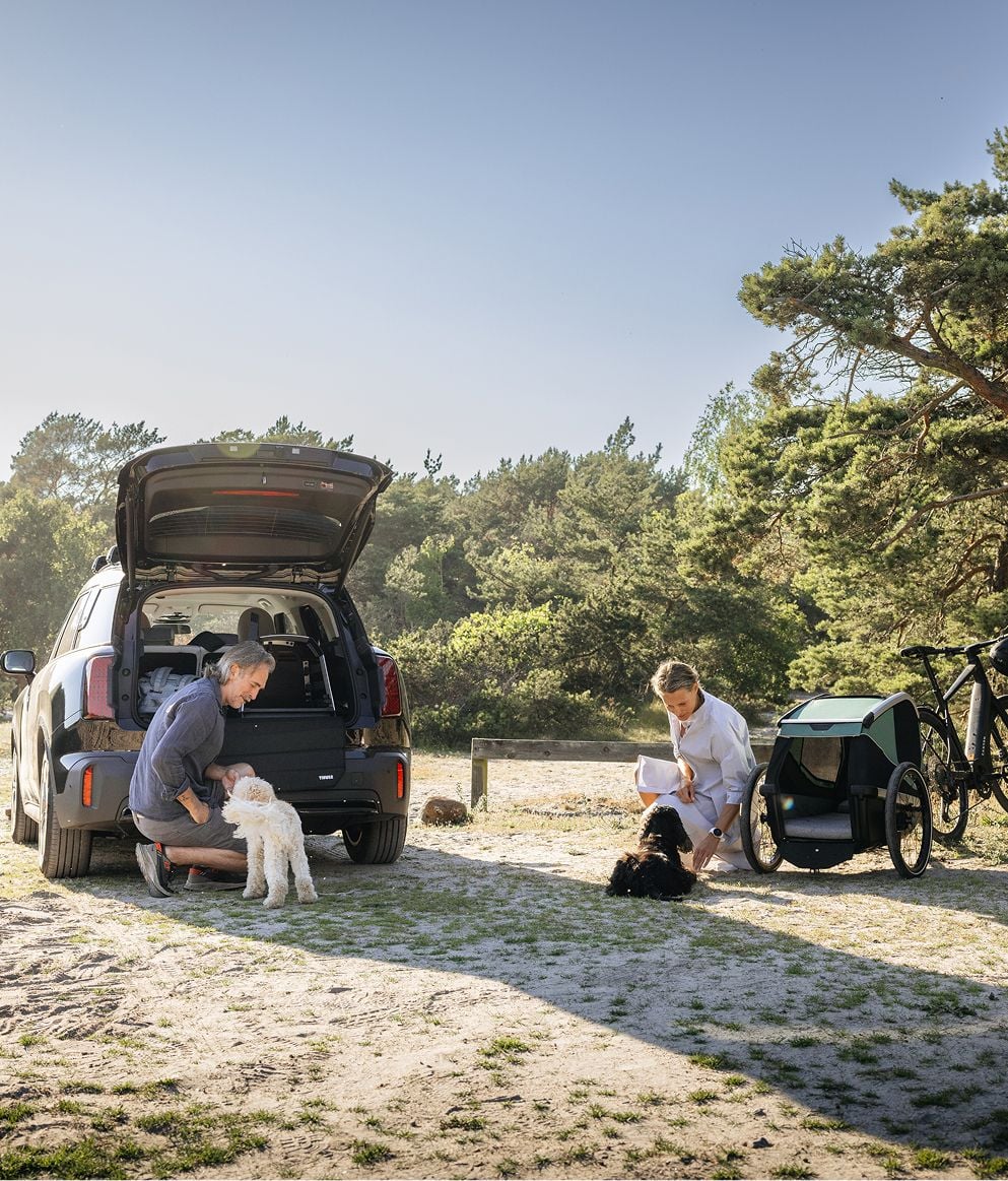 Two persons with dogs beside a vehicle equipped for dog travel, with a dog trailer attached to a bicycle nearby.