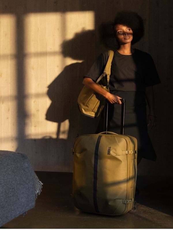 Imagem de uma pessoa de pé com sacos Thule Aion, banhada numa luz cálida e natural, incluindo um saco com rodas e um saco de compras.