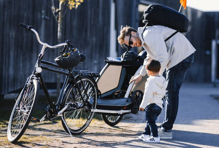 A father helps his toddler get in the Thule Courier baby bike trailer that is attached to his black city bike.