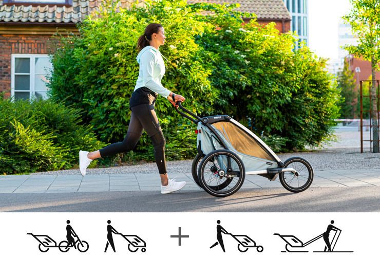 A woman in sportswear jogs down a residential street with a blue Thule Chariot Cross jogging bicycle trailer.