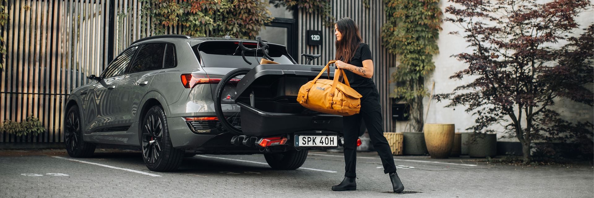 Woman loading a yellow bag into a rear cargo carrier on a silver SUV parked outside a modern, ivy-covered building.