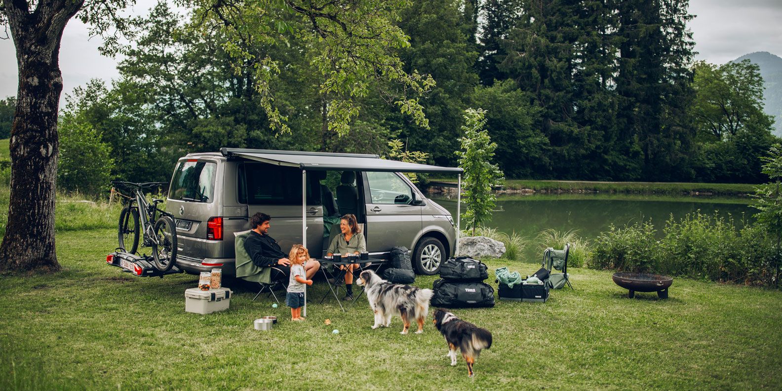 A compact van is parked in the grass and a family sits under the Thule Sidehill compact van awning.