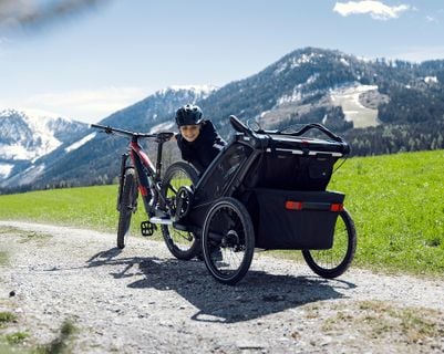 跪著的人坐在一輛帶有自行車拖車的登山車旁，行駛在碎石路上，地點是山區景觀。