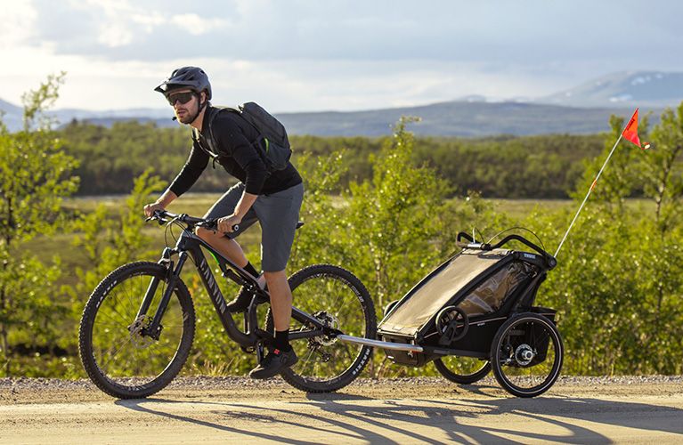 In an expansive grassland a man goes biking with a black Thule Chariot Sport child bike trailer.