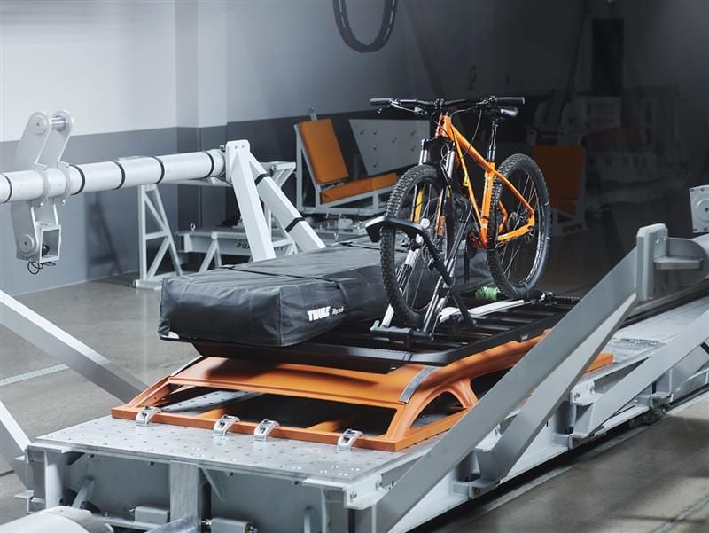 Una struttura di crash test con una slitta di prova con un sistema di trasporto per bici e carichi Thule dal montaggio posteriore. Una mountain bike di colore arancione brillante è fissata saldamente al sistema di portaggio insieme a una borsa cargo nera con marchio Thule.