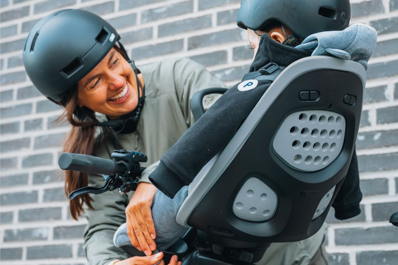 Gros plan d’un adulte attachant la sangle de pied d’un enfant pendant que l’enfant est assis sur un siège arrière de vélo, tous deux portant un casque.