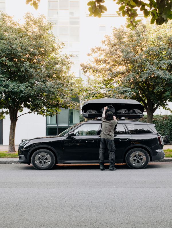 En person laster utstyr inn i en svart Thule Force 3-takboks montert på en svart bil som er parkert under løvtrær i urbane omgivelser