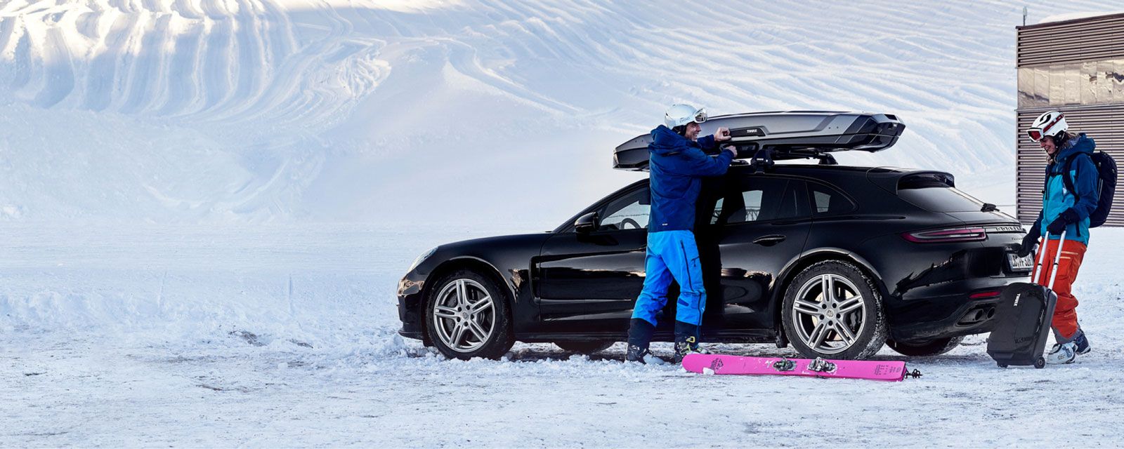 In the snow, one skier opens the Thule roof box while the other rolls a Thule wheeled duffel bag.