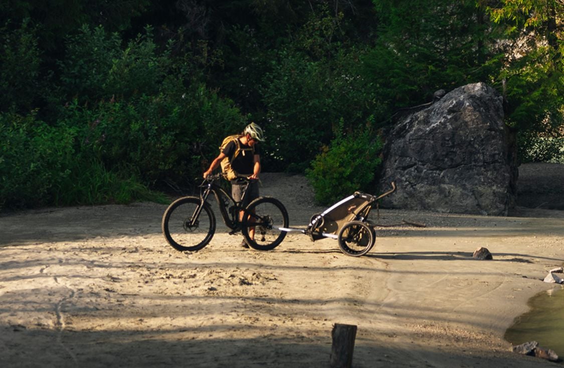 Person standing with a bicycle, pulling a bike trailer along a sandy riverbank surrounded by trees and rocks.
