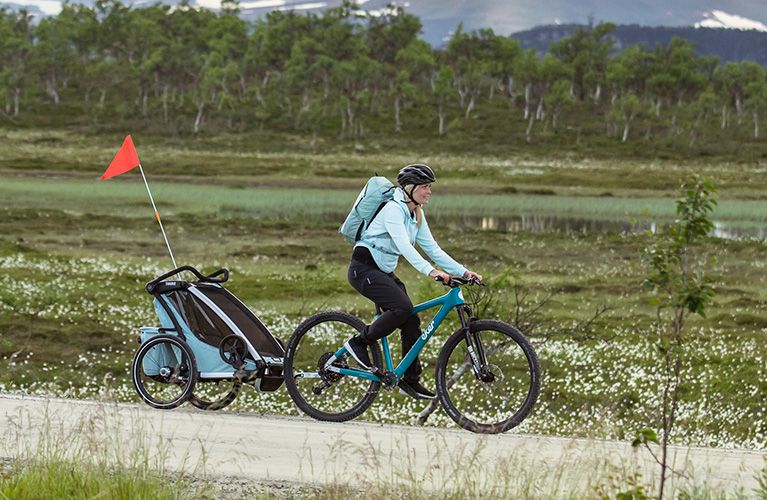 Kobieta jedzie przez pole z dziecięcą przyczepką rowerową Thule Chariot Cross.