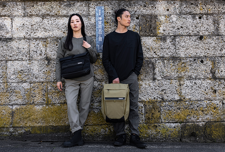 Two people stand with their back to a mossy wall holding the Thule Paramount crossbody and backpack.