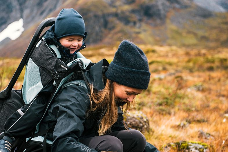 A mother leans down in a field with her child in a Thule child carrier backpack. 