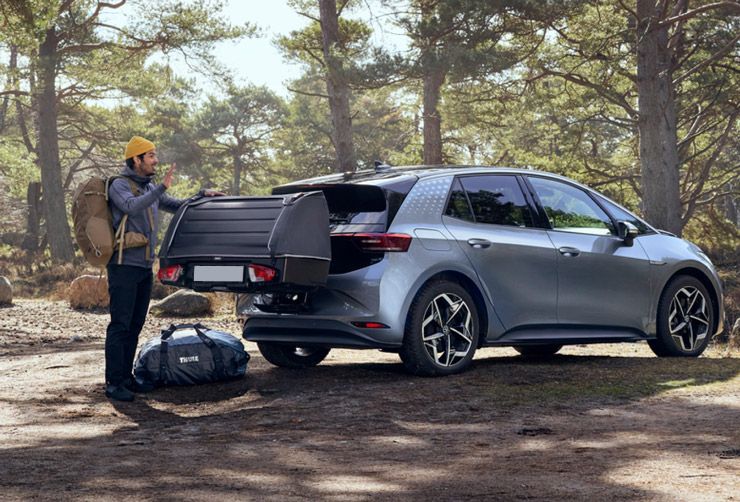 A car is parked in forest with a Thule rear cargo carrier and a man with a backpack stands beside it.