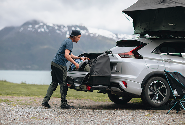 A man loading off bags from a Thule Arcos cargo box that is mounted on the back of a car, parked in front of lake with surrounding mountains. 