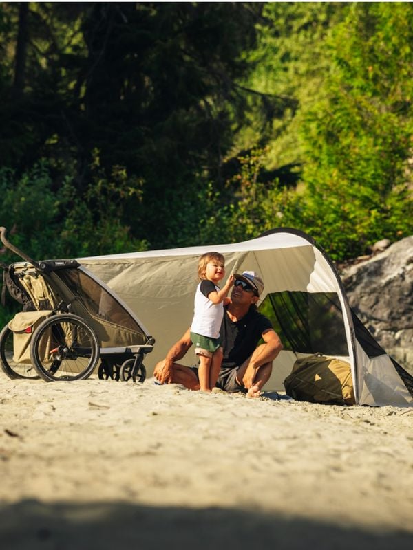 Eltern und Kind entspannen an einem Sandstrand unter einer Plane des Typs Thule Sun & Wind Tarp. In der Nähe Kinderwagen und Fahrrad, im Hintergrund Wald.