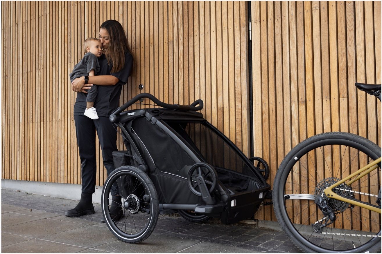 A woman holds her baby leaning against a wooden wall next to a bike trailer jogging stroller.