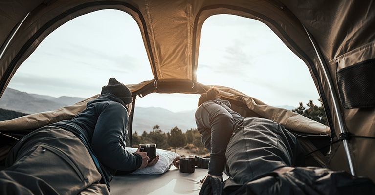 A couple sit inside a roof top tent looking outside the windows.