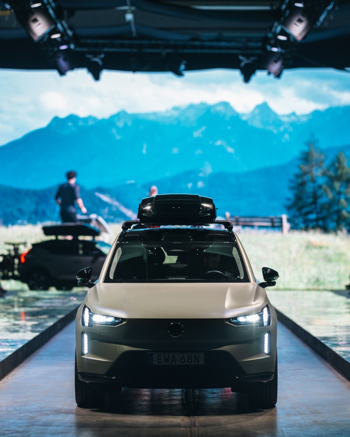 A car with a roof box driving on a runway in front of a large screen showing a mountain landscape.