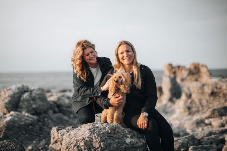 Two women are sitting with their dog on a rocky beach.