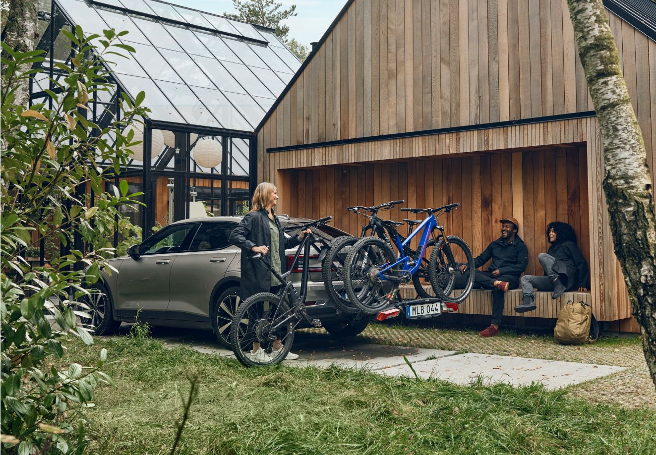 En person laster en cykel på en cykelholder bag på en bil, mens to personer sidder smilende på en træbænk.