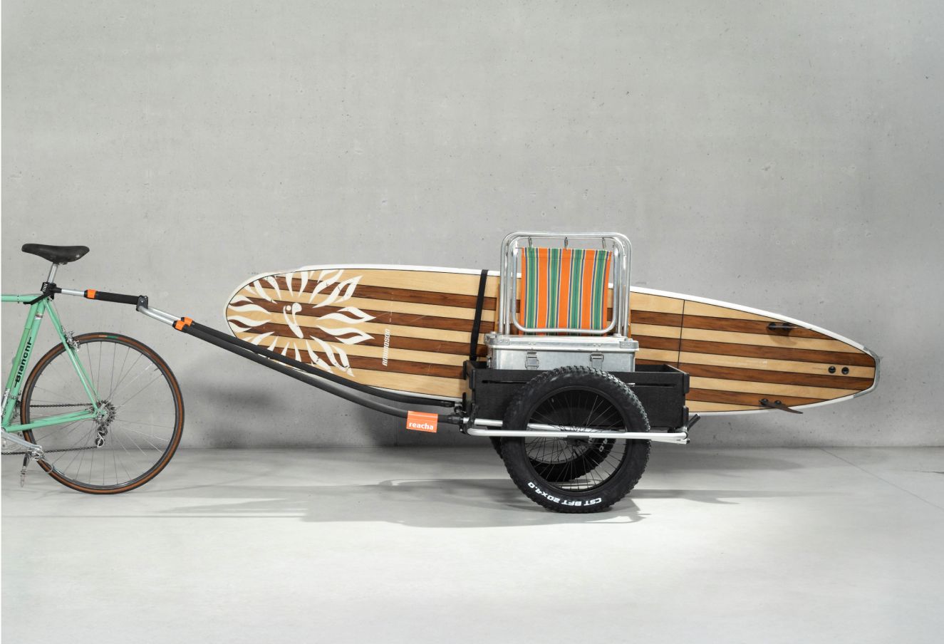 Una bicicleta verde con un remolque de bicicleta Reacha Sport Beach conectado en el que se transporta una tabla de surf a rayas, todo mostrado en un estudio minimalista.