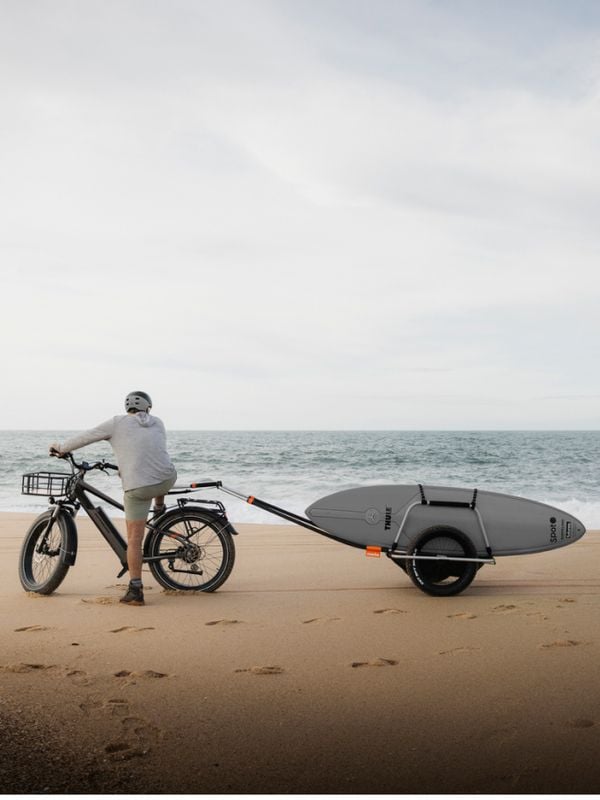 Uma pessoa com uma bicicleta e um atrelado Reacha numa praia, a transportar uma prancha de surf com o mar em plano de fundo
