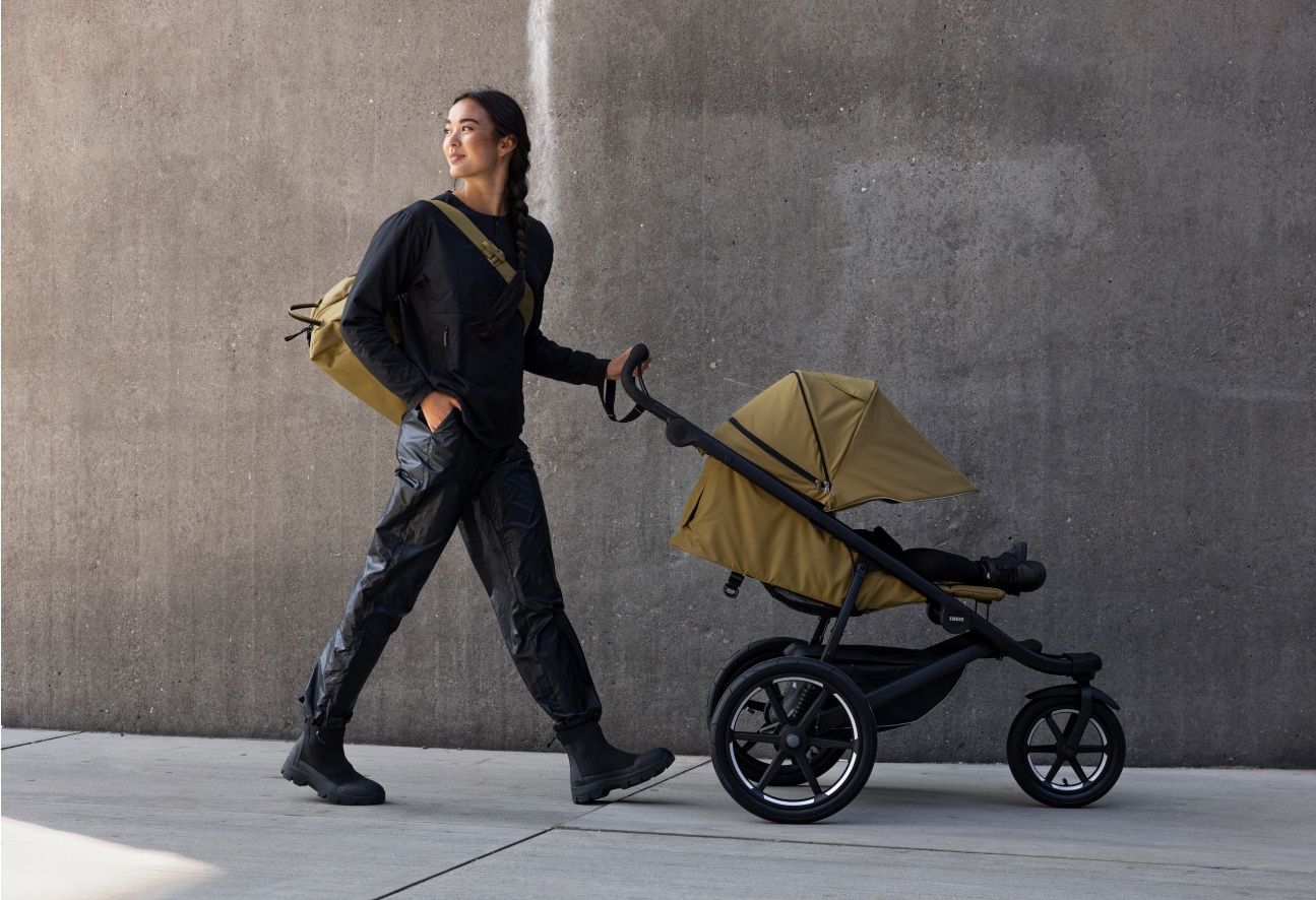 A woman wearing a Thule backpack looks behind her as she pushes a Thule stroller.