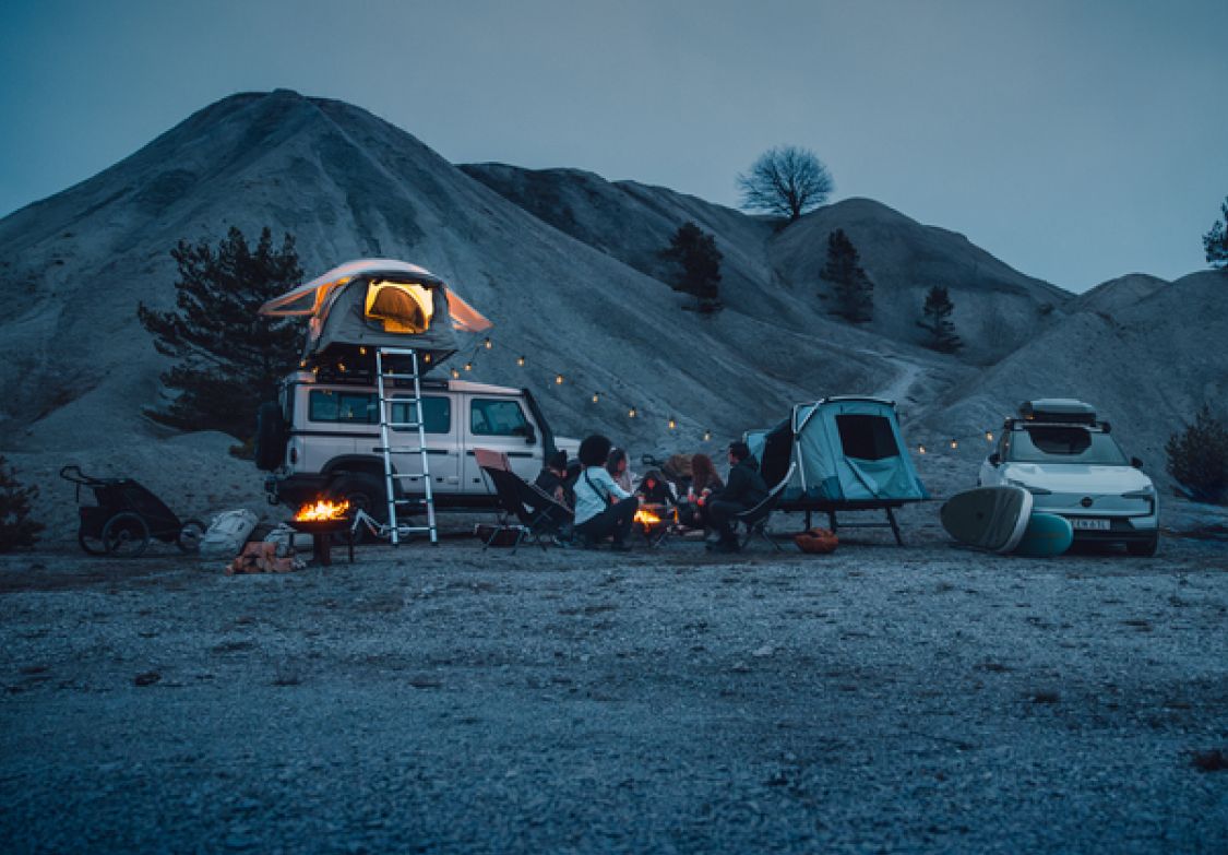 Inimesed istuvad õhtuhämaruses lõkke ümber kahe sõiduki kõrval, mis on varustatud Thule telkide ja toodetega.