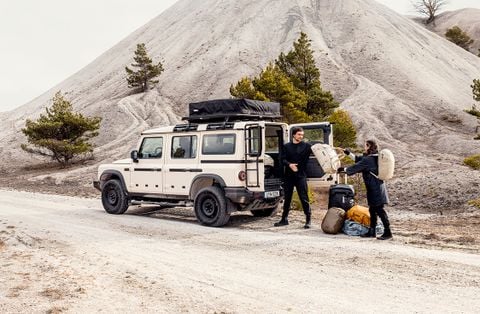 Two people unloading gear next to a rugged off-road vehicle equipped with a rooftop tent, parked in a remote, rocky landscape.