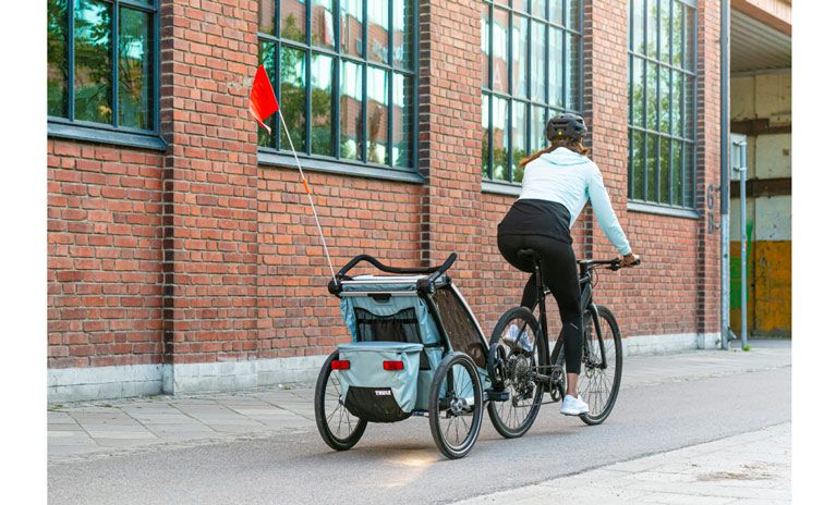 A woman bikes by a brick building with the child bike trailer Thule Chariot Cross.