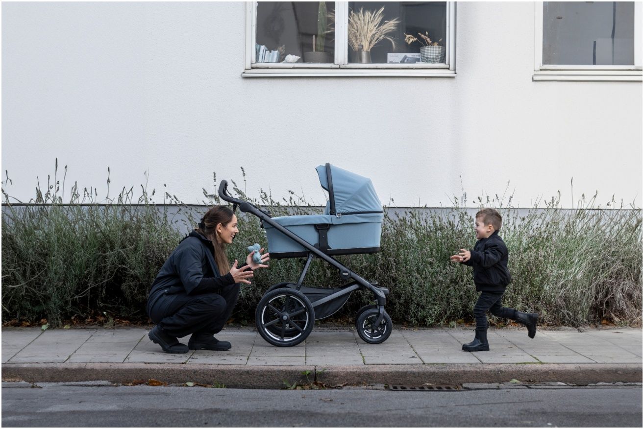 In a park a man leans down and tends to his kids inside a Thule double stroller.