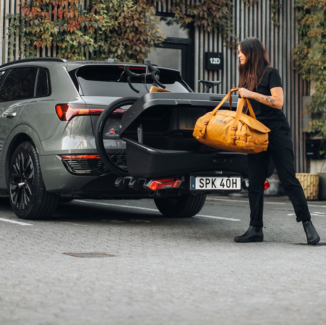 Woman loading a yellow bag into a rear cargo carrier on a silver SUV parked outside a modern, ivy-covered building.