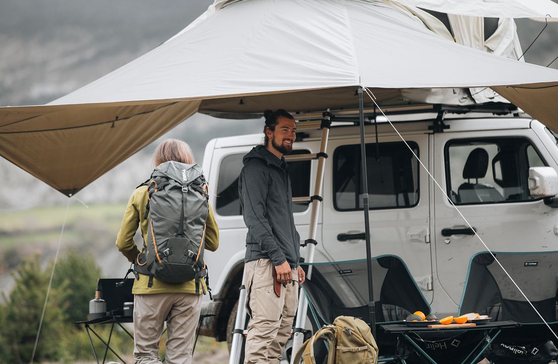 Two people standing under a mounted awning attached to a van equipped with Thule gear, including a rooftop tent and ladder.