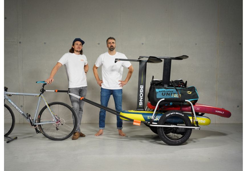 Dwóch mężczyzn pozuje w studio z przyczepką rowerową Reacha Sport Beach przymocowaną do roweru, na przyczepce deski surfingowe i sprzęt rekreacyjny.