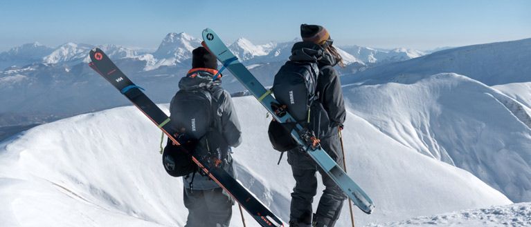 Alice Linari and Lorenzo Alesi stand on a snowy mountain looking at the view with skis and Thule ski backpacks.