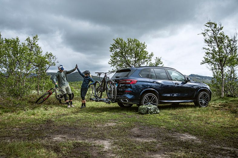 Une auto est stationnée sur la pelouse et un couple se tape dans les mains, ils sont appuyés contre leurs vélos sur un support pour vélos sur attelage.