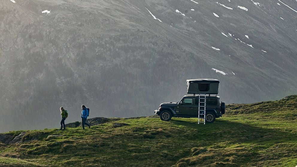 Zwei Personen wandern auf einem grasbewachsenen Weg neben einem Fahrzeug mit einem Dachzelt mit stabiler Außenhülle auf den Dachträgern.