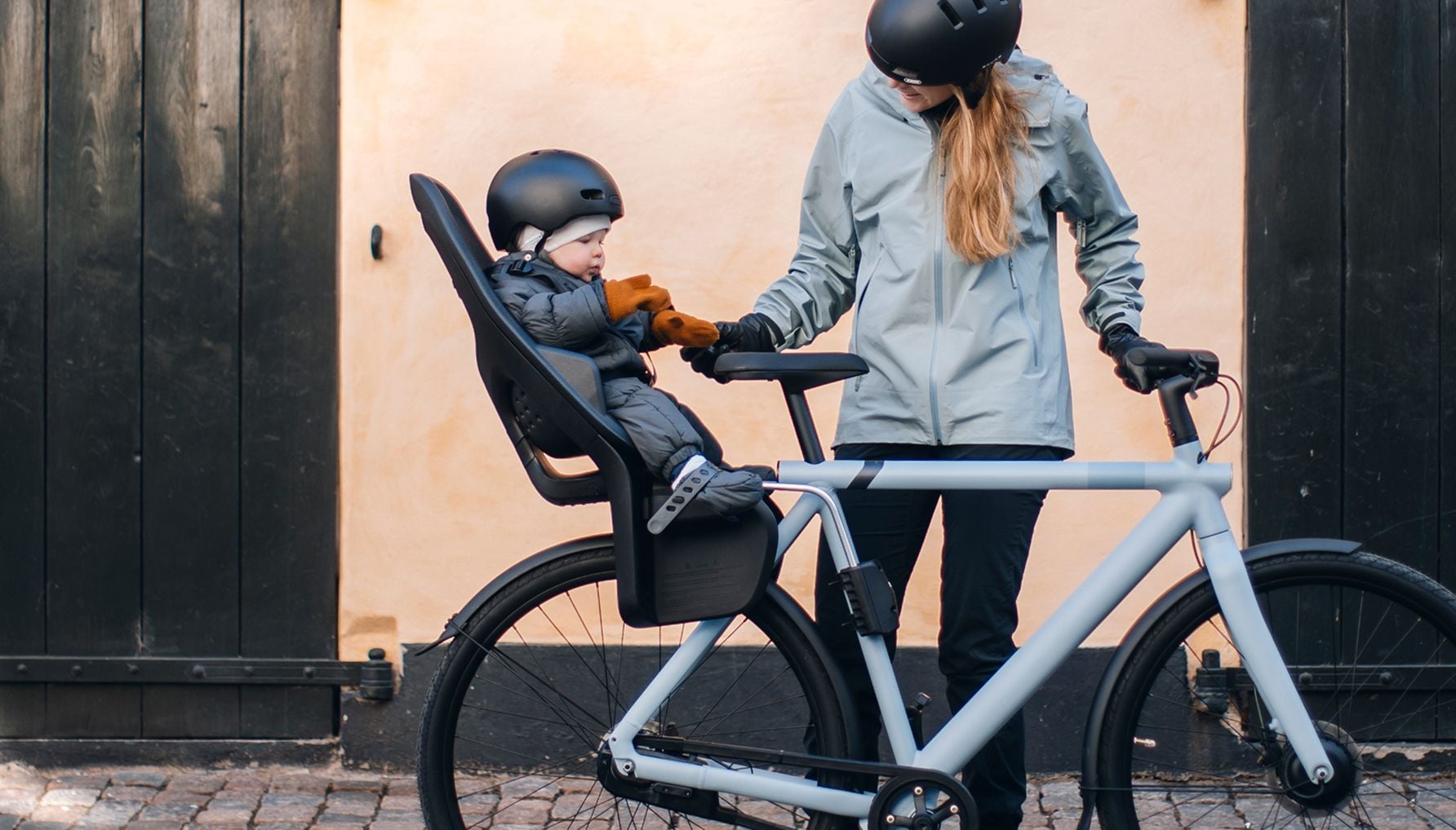 Adult standing with a bicycle, interacting with a child seated in a Thule Yepp 2 maxi frame-mounted bike seat, both wearing helmets in front of a building wall.