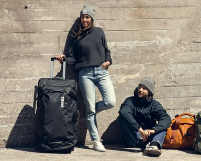 Två personer lutar sig mot en betongvägg och håller i stora resväskor.